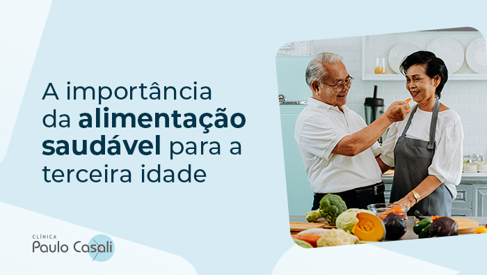 A imagem mostra um casal de idosos cozinhando. Enquanto a mulher corta legumes, o homem dá um pedaço de comida na boca dela.